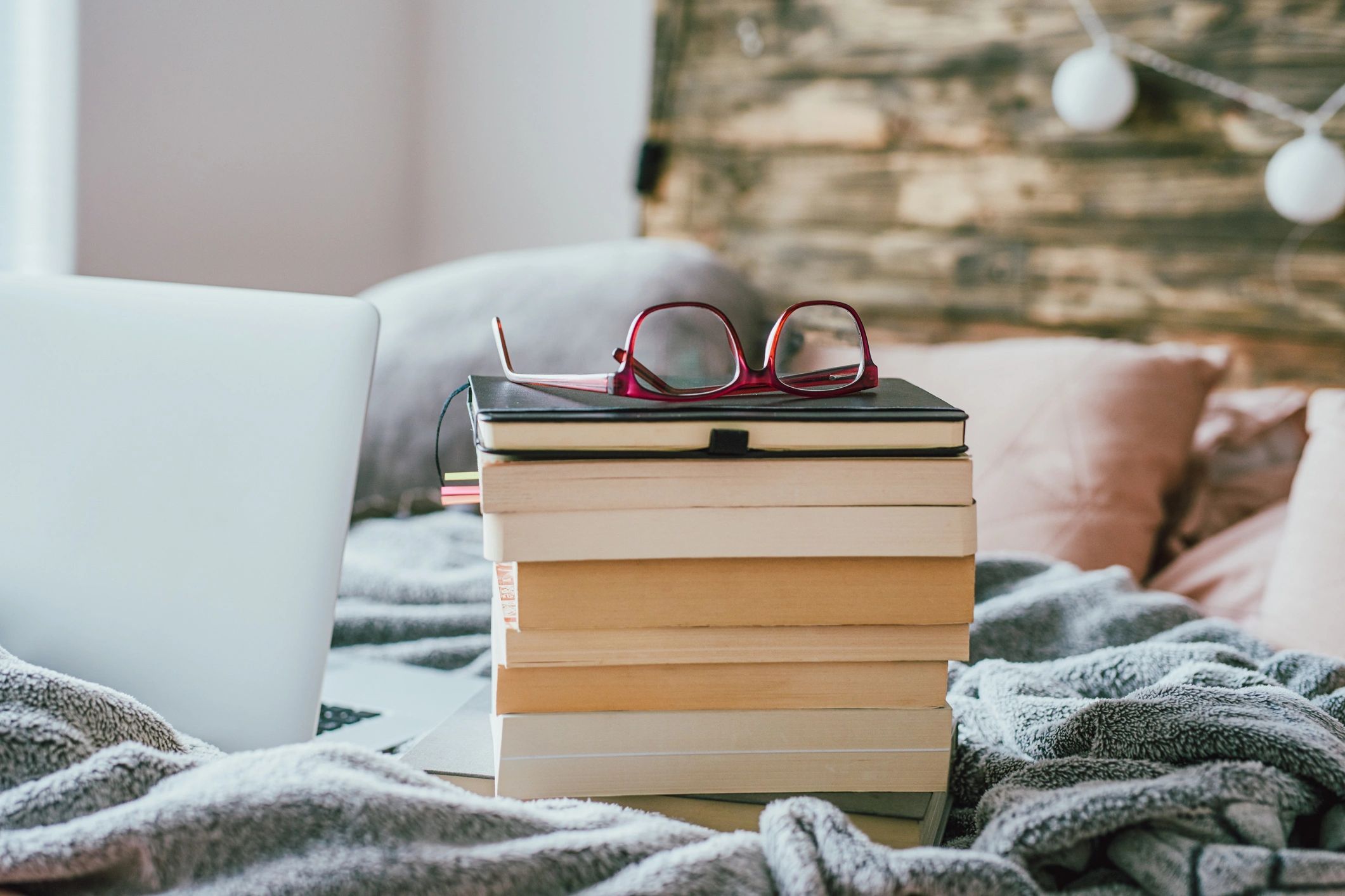 Stack of books and glasses on a bed next to a laptop